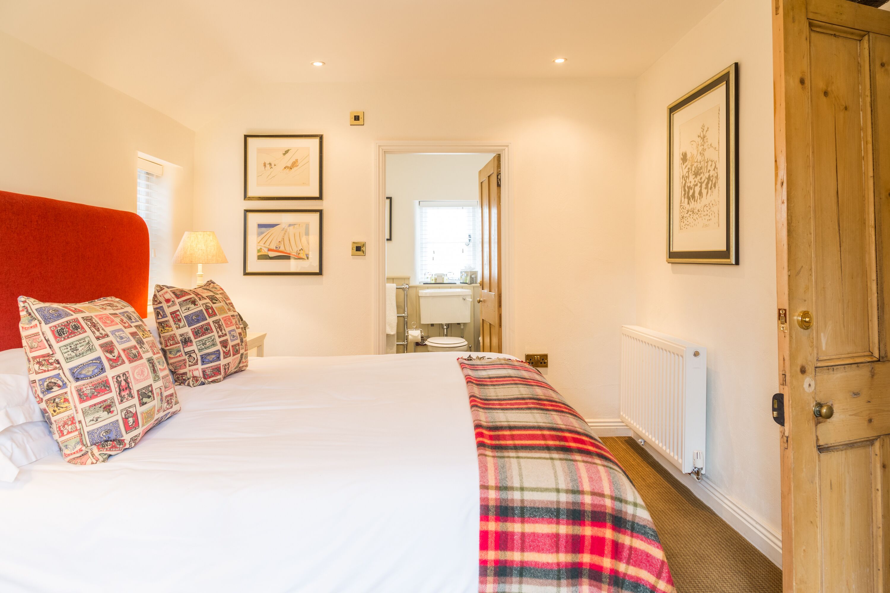 A hotel room with a large bed, a red headboard, a plaid blanket, and two pillows. There is a door on the right and a bathroom visible through an open door in the background.