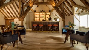 A bar area with wooden beams, a glass table, and bar stools. There is a shelf with bottles and a sign that reads "THE DAY". - Old Swan & Minster Mill Hotel (Witney)