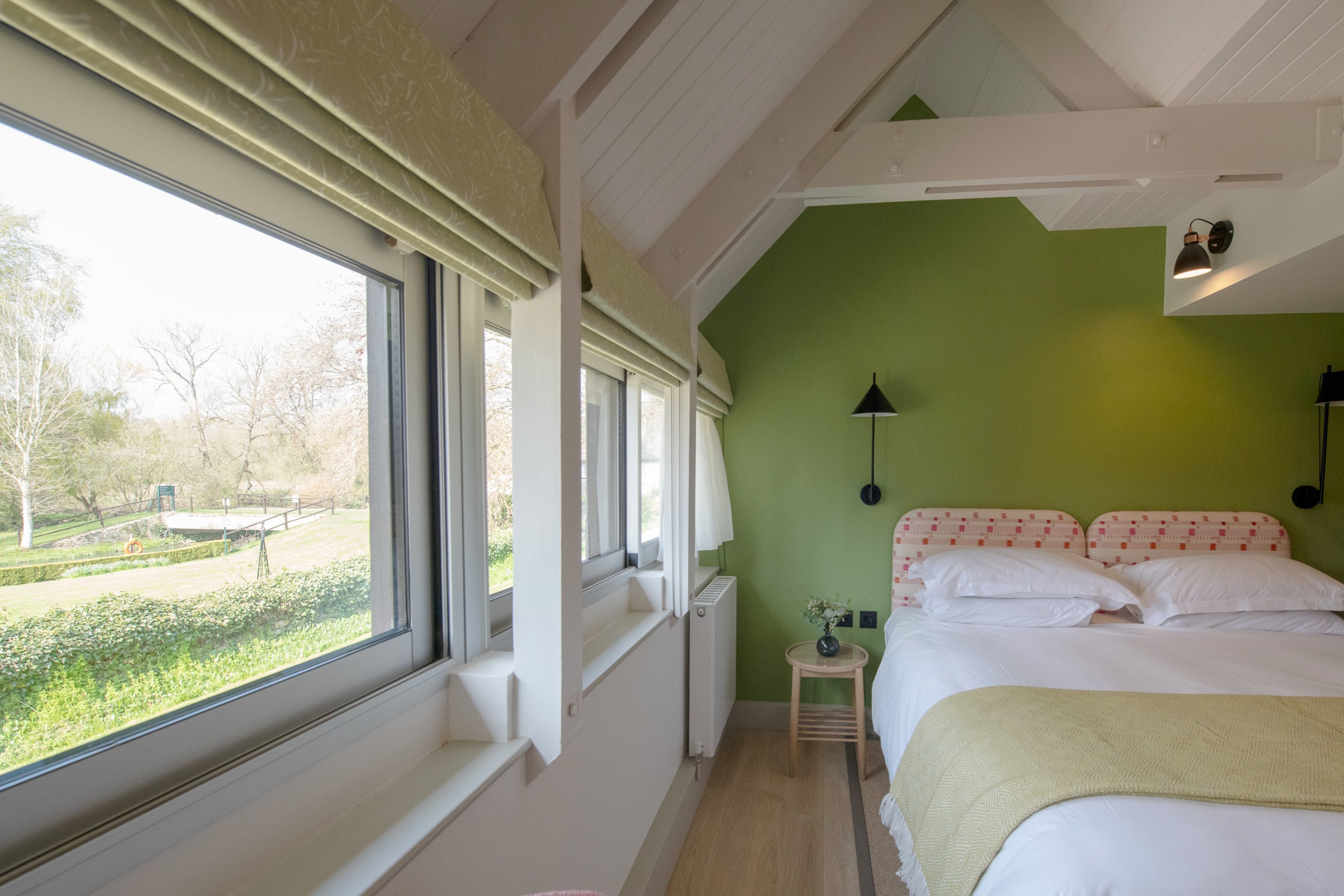 A bedroom with a large window, a bed with white bedding, a small bedside table, and a green wall.