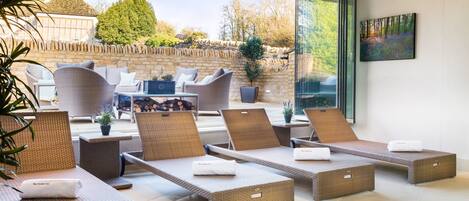 A sunlit outdoor lounge area with wicker loungers, a stone wall, and a large window offering a view of the surrounding landscape.