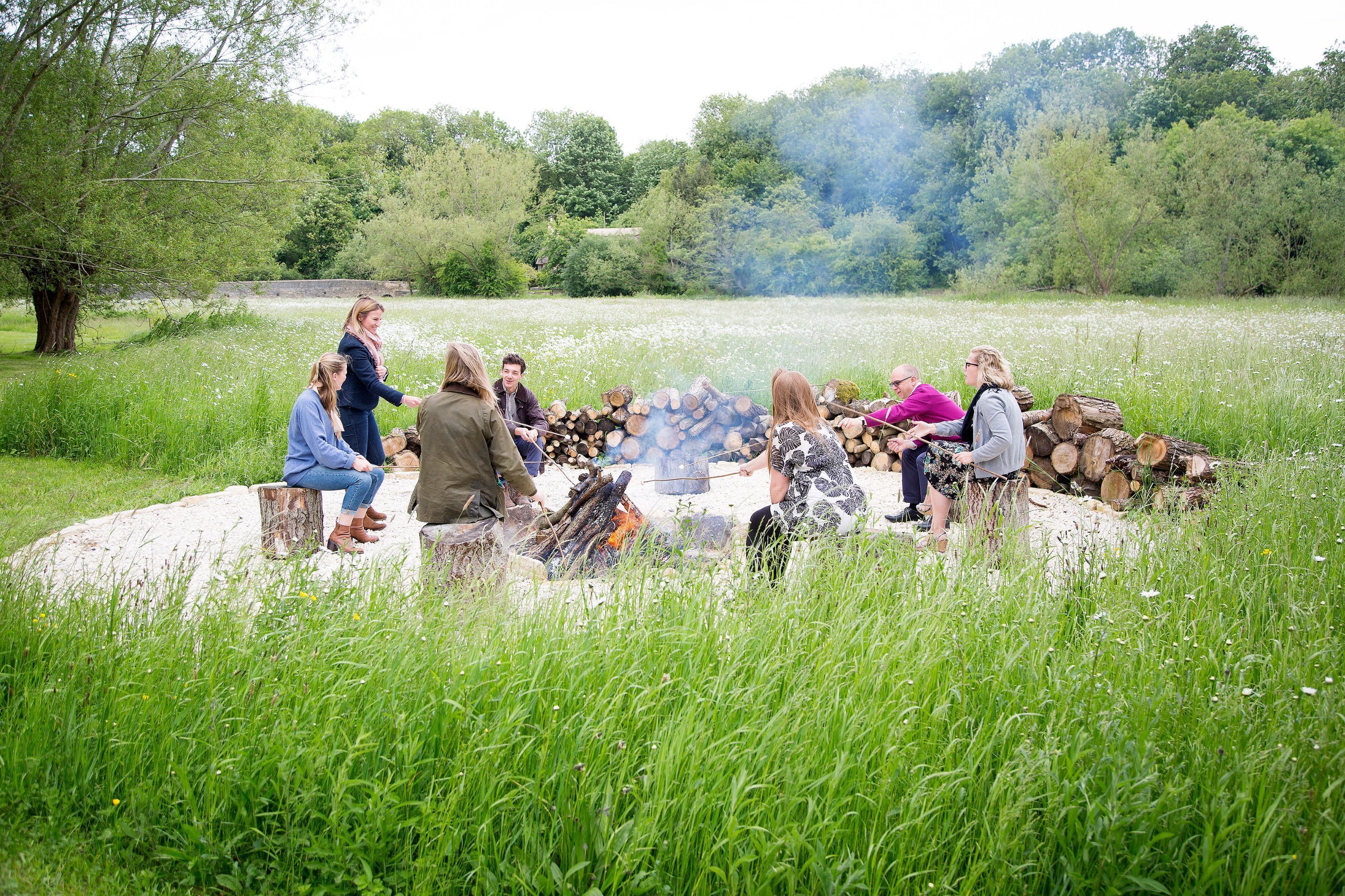 A group of people gathered around a campfire in a grassy field with trees in the background.