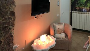 Een gezellige woonkamer met een aan de muur bevestigde televisie, een klein tafeltje met kaarsen, een rieten fauteuil en een stenen muur.