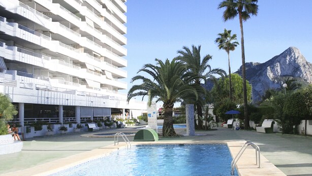 A swimming pool area with a large building in the background, palm trees, and a mountain. - Apartamentos Esmeralda (Calpe)