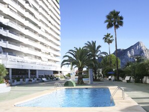 A swimming pool area with a large building in the background, palm trees, and a mountain. - Apartamentos Esmeralda (Calpe)