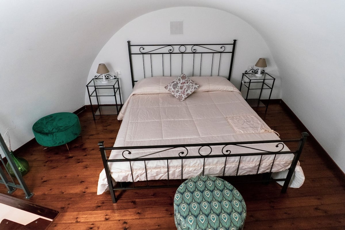 A single bed with a headboard, two bedside tables with lamps, a green ottoman, and a patterned ottoman.