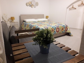 Un dormitorio con cama, mesitas de noche, un taburete, una silla, un banco y una planta en maceta.