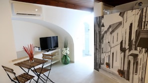 A modern living room with a TV, a dining table with chairs, a vase with flowers, and a wall with a black and white cityscape mural. - Il Vicolo B&B (Putignano)