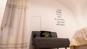 A cozy room with a grey sofa, a decorative pillow, a wall with a quote, and a partially visible bed. - Il Vicolo B&B (Putignano)
