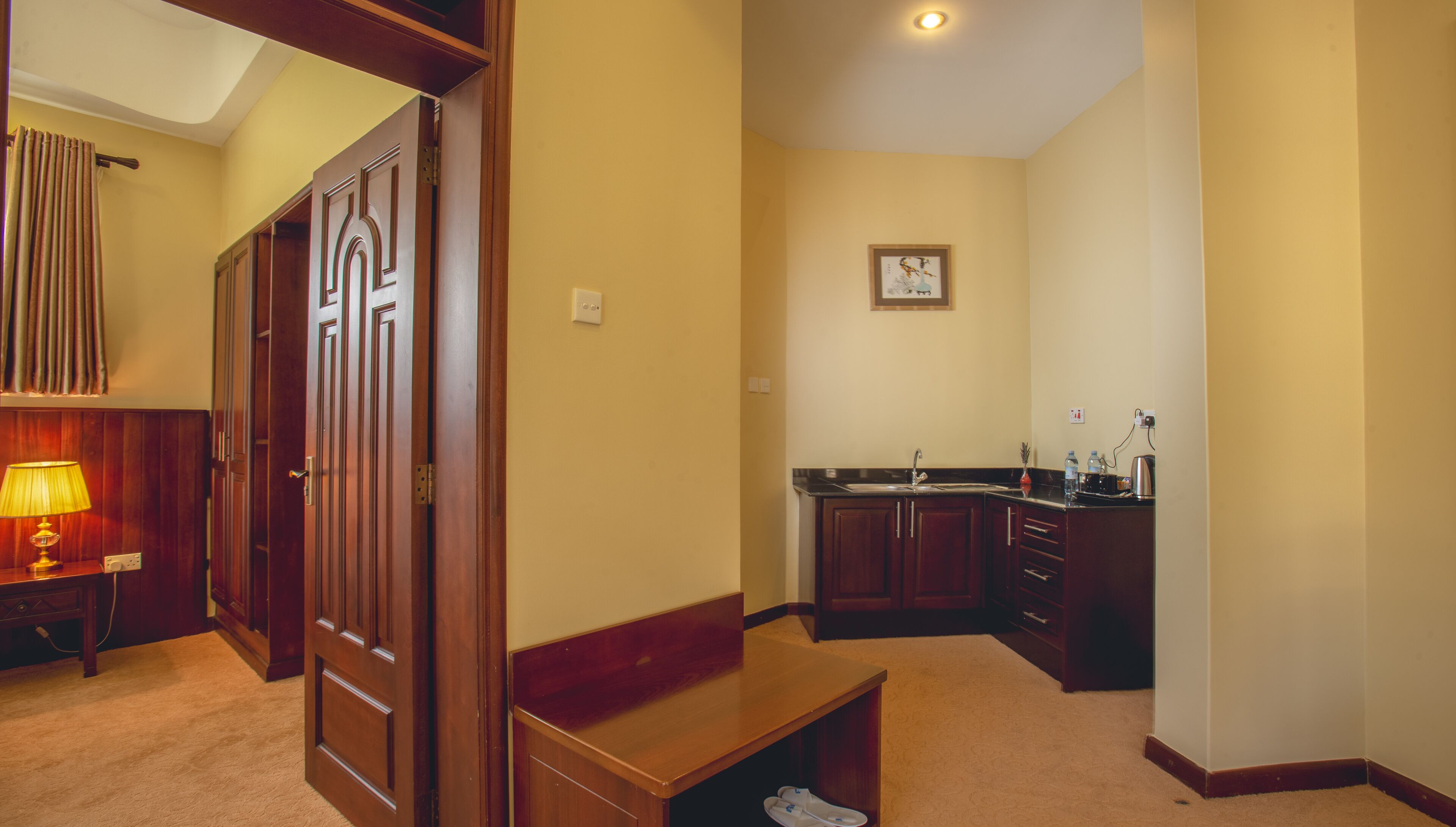 A hotel room with a wooden wardrobe, a bench, a desk with a lamp, a kitchen area with a sink and cabinets, and a framed picture on the wall.