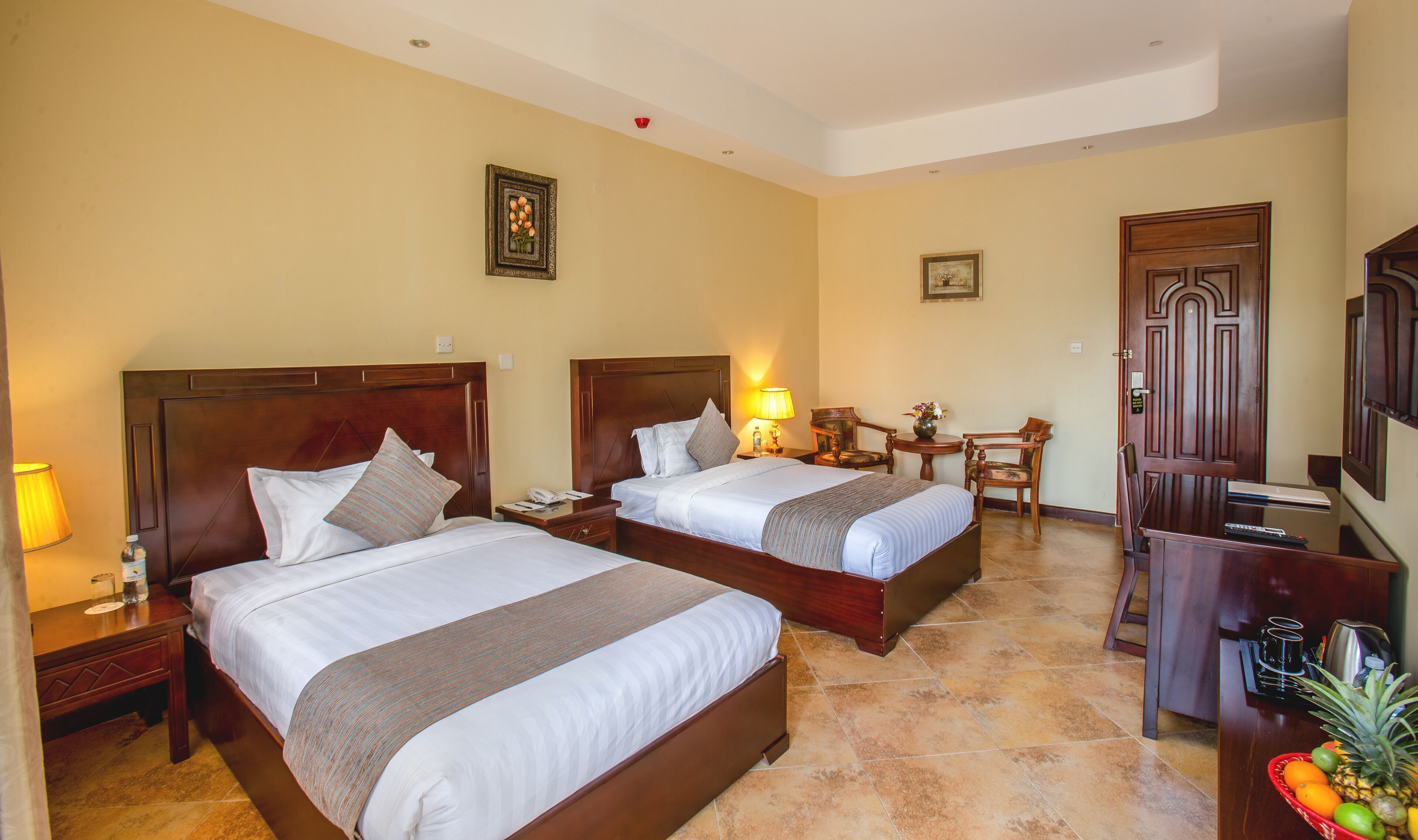 A hotel room with two beds, a desk, a chair, a small table with a lamp, and a door.