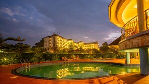 A swimming pool with a reflection of a building, surrounded by greenery and a well-lit balcony. - Nyaika Hotel (Fort Portal)