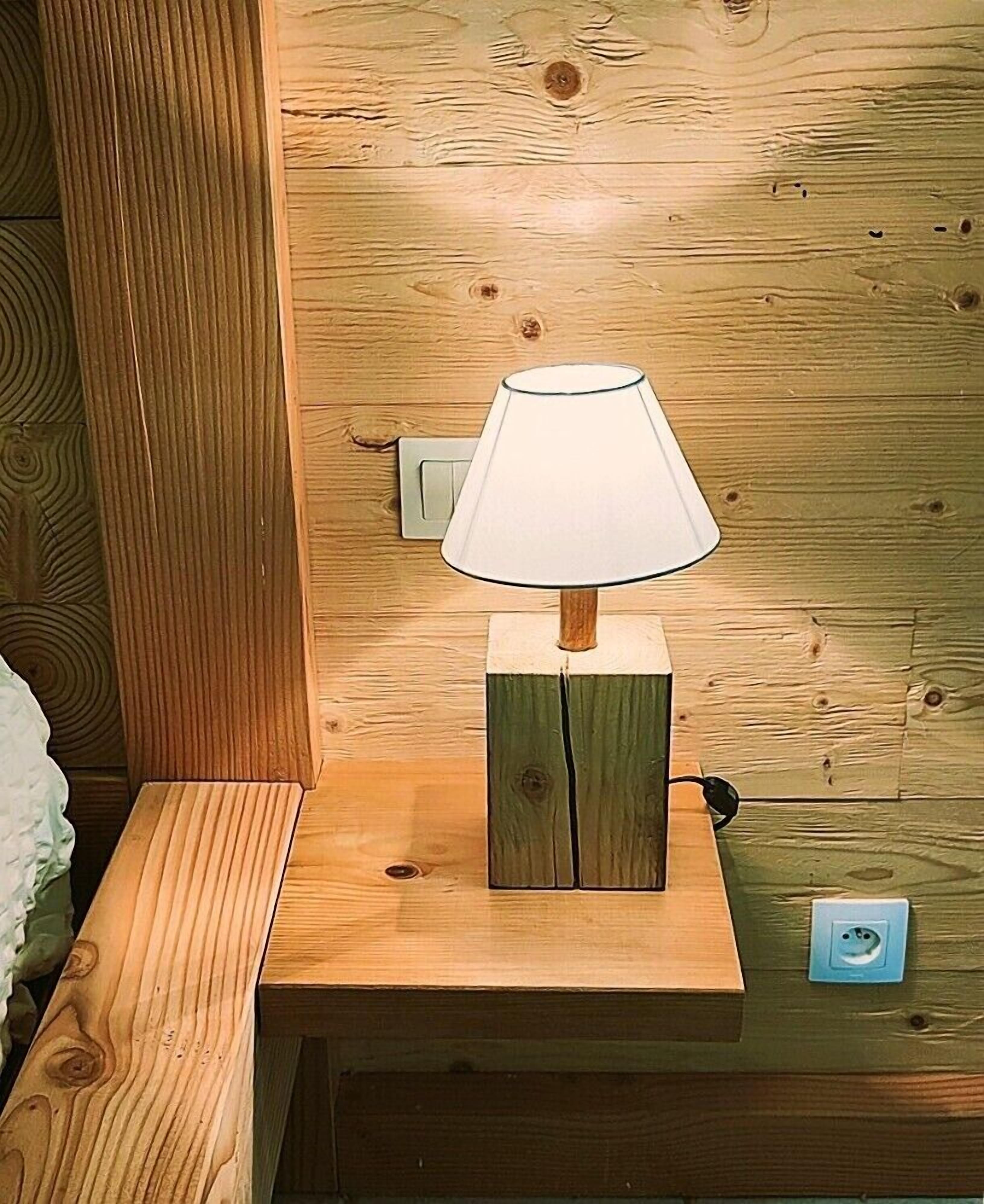 Une table de chevet en bois, avec une lampe, une prise de courant et un interrupteur au mur.