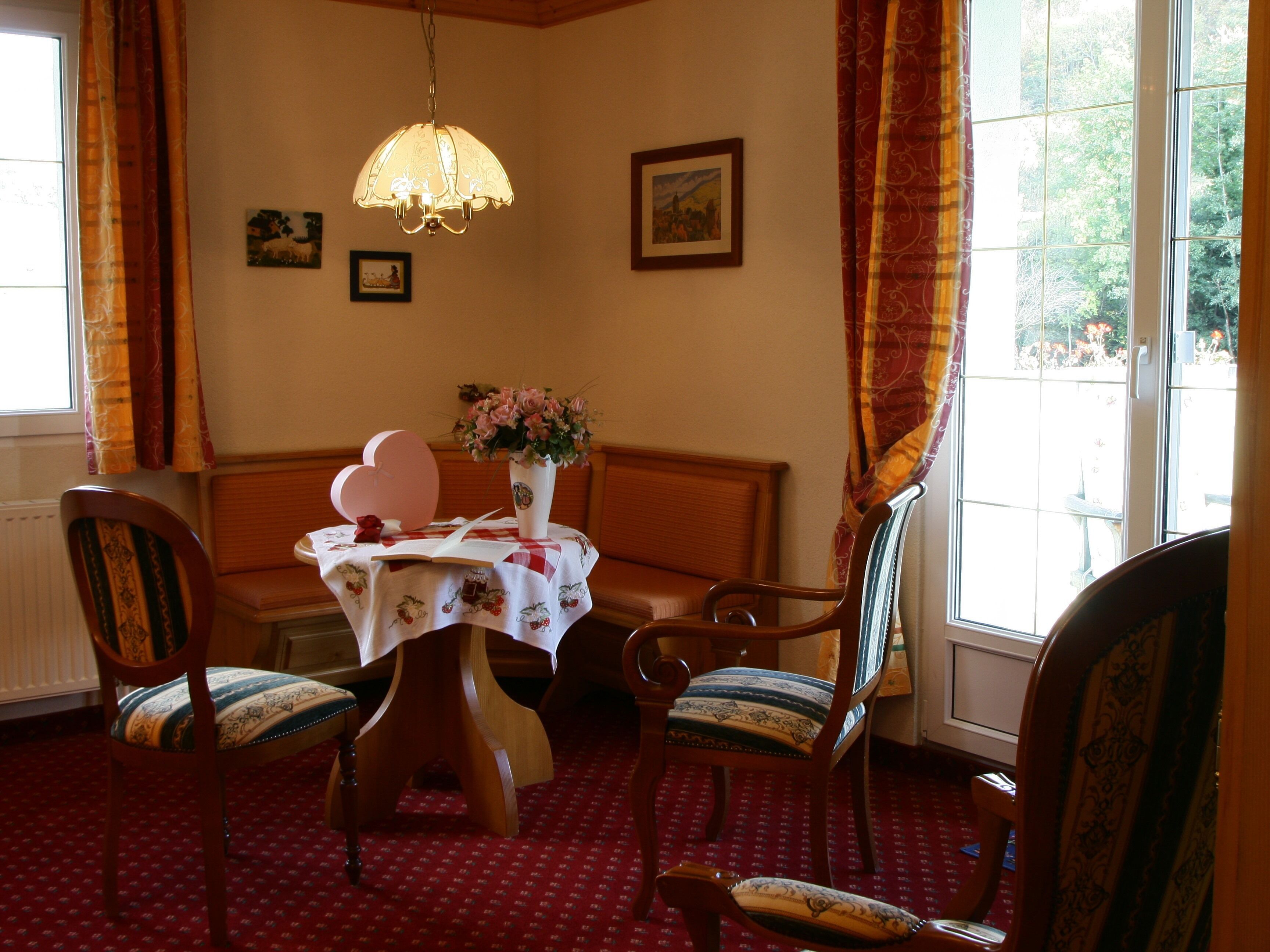 A dining area with a table set for two, surrounded by wooden chairs with patterned cushions. A chandelier hangs from the ceiling, and there are two framed pictures on the wall.