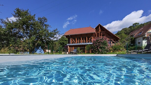 Une piscine à l’eau claire, entourée d’un bâtiment en bois avec un toit rouge, et une végétation luxuriante en arrière-plan.
