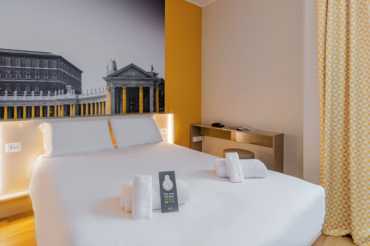 Habitación de hotel con una cama grande, una mesita de noche, un escritorio y un mural de un edificio en la pared.