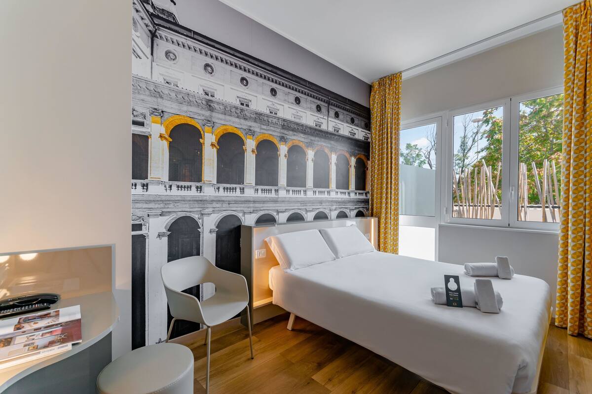 Habitación de hotel con cama, escritorio, silla y un amplio ventanal con cortinas.