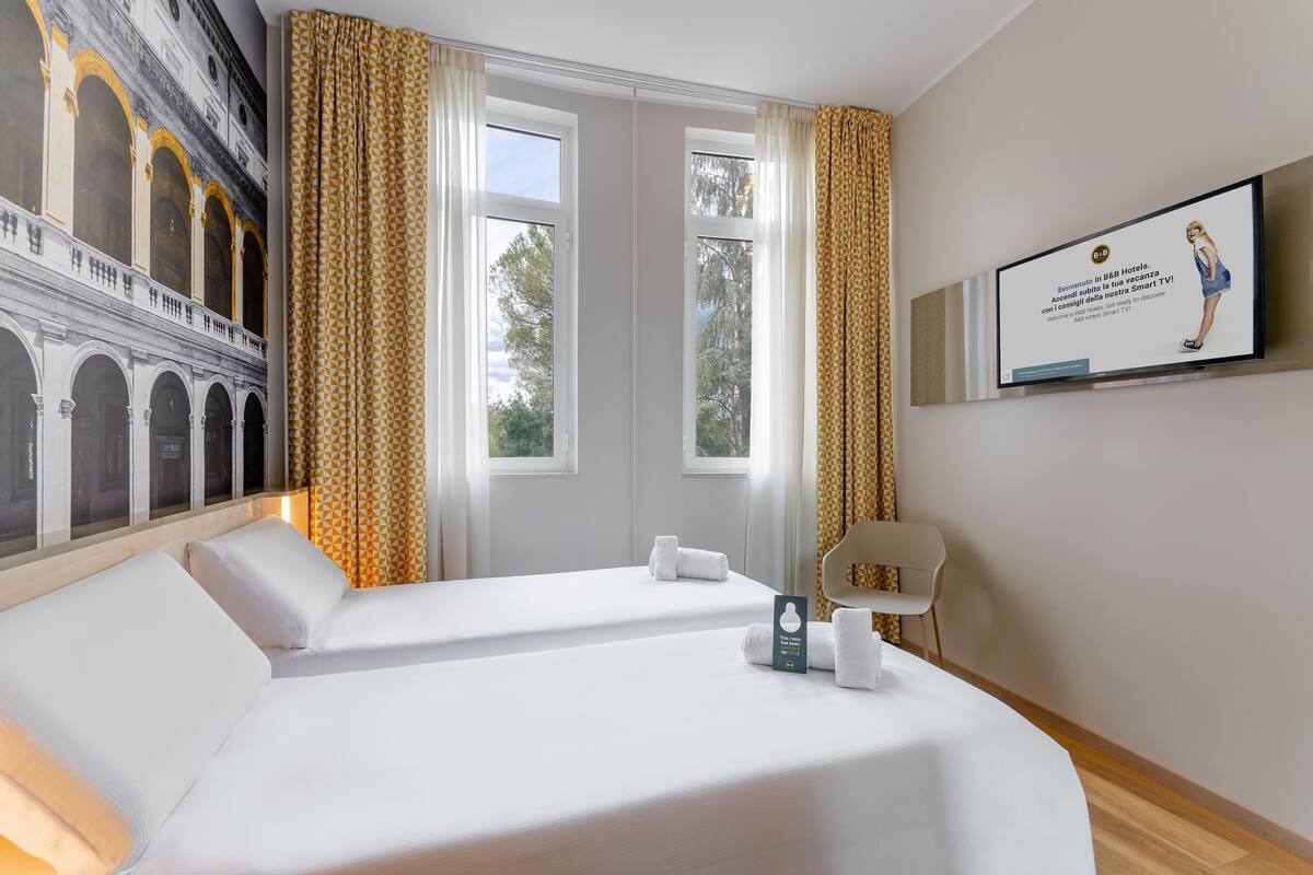 Habitación de hotel con dos camas, televisión y vistas a un edificio a través de las ventanas.