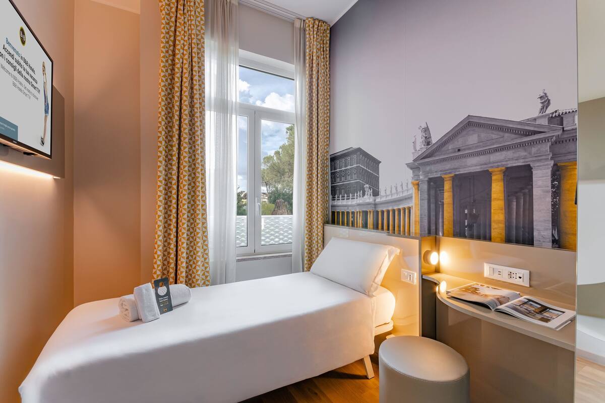 Una habitación de hotel con cama, una mesita con una revista, una ventana con cortinas y un mural en la pared.