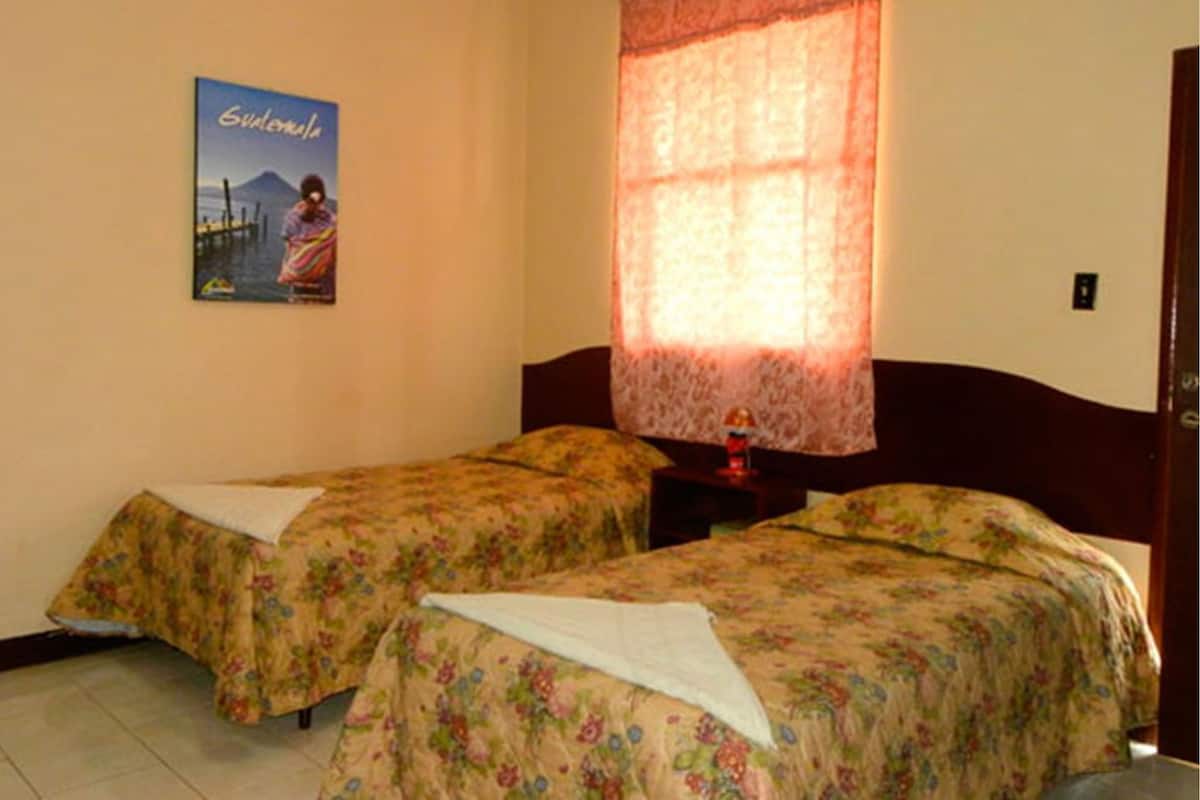 A room with two beds, each with floral bedding, a poster on the wall, and a window with a lace curtain.
