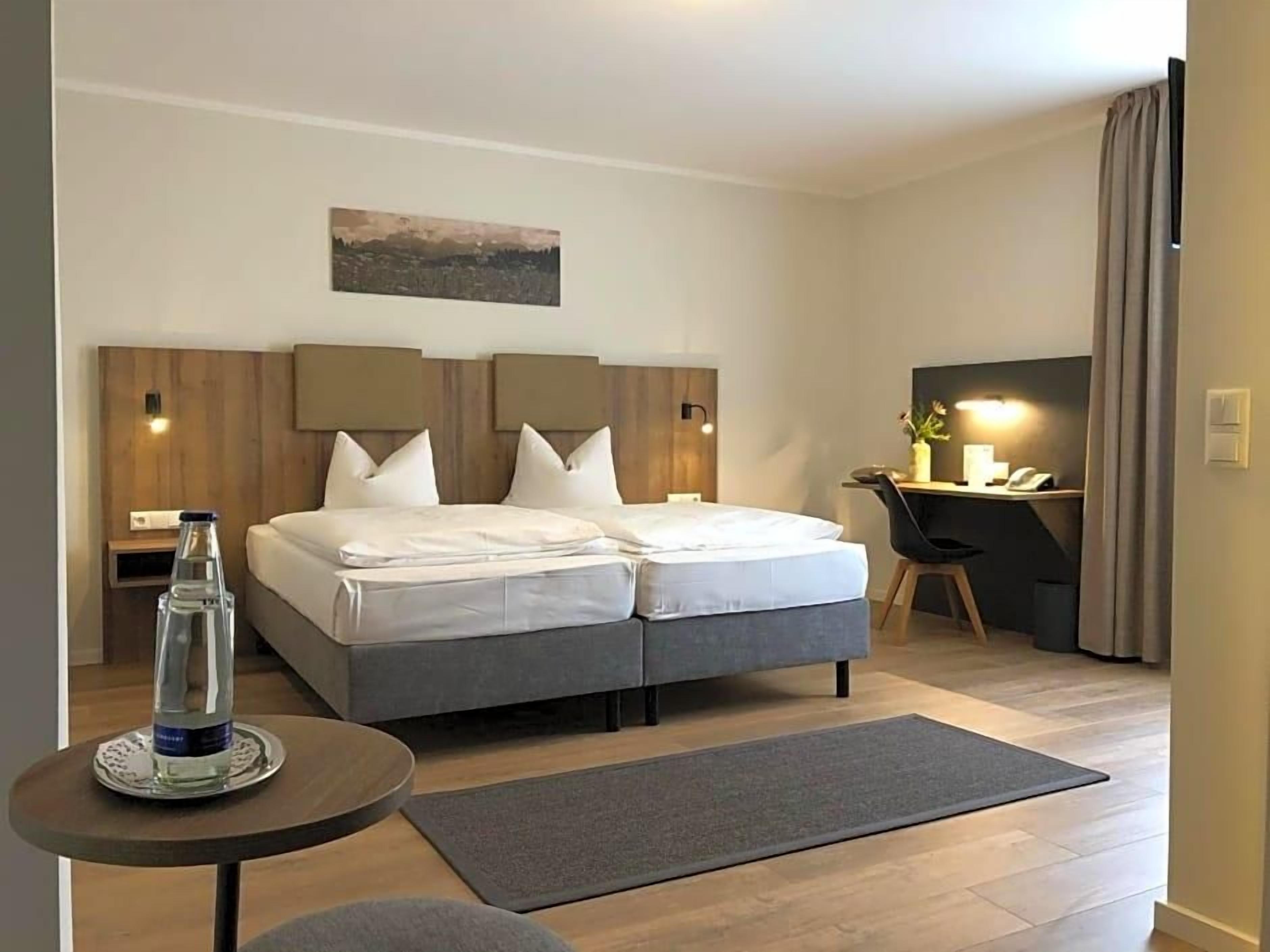 A hotel room with a double bed, a desk, a chair, a small table with a bottle, and a painting on the wall.