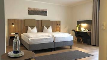 A hotel room with a double bed, a desk, a chair, a small table with a bottle, and a painting on the wall.