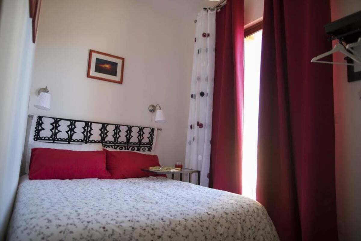 A bedroom with a bed, red pillows, a small table with a book and a glass, a wall-mounted lamp, and a framed picture.