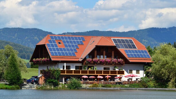 A building with a red roof and solar panels, located near a body of water with mountains in the background. - Gasthof Fischerwirt (Neumarkt in Steiermark)