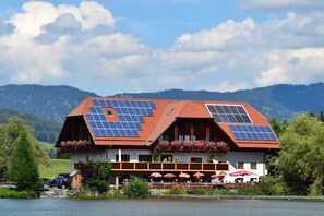 A building with a red roof and solar panels, located near a body of water with mountains in the background. - Gasthof Fischerwirt (Neumarkt in Steiermark)