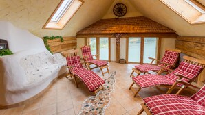 A cozy attic room with a sloped ceiling, wooden walls, and a large window overlooking a river. There are several wooden chairs with red and white checkered cushions, a small fireplace, and a mosaic-tiled floor. - Gasthof Fischerwirt (Neumarkt in Steiermark)