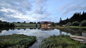 A lakeside view with a building reflecting on the water, surrounded by greenery and trees. - Gasthof Fischerwirt (Neumarkt in Steiermark)