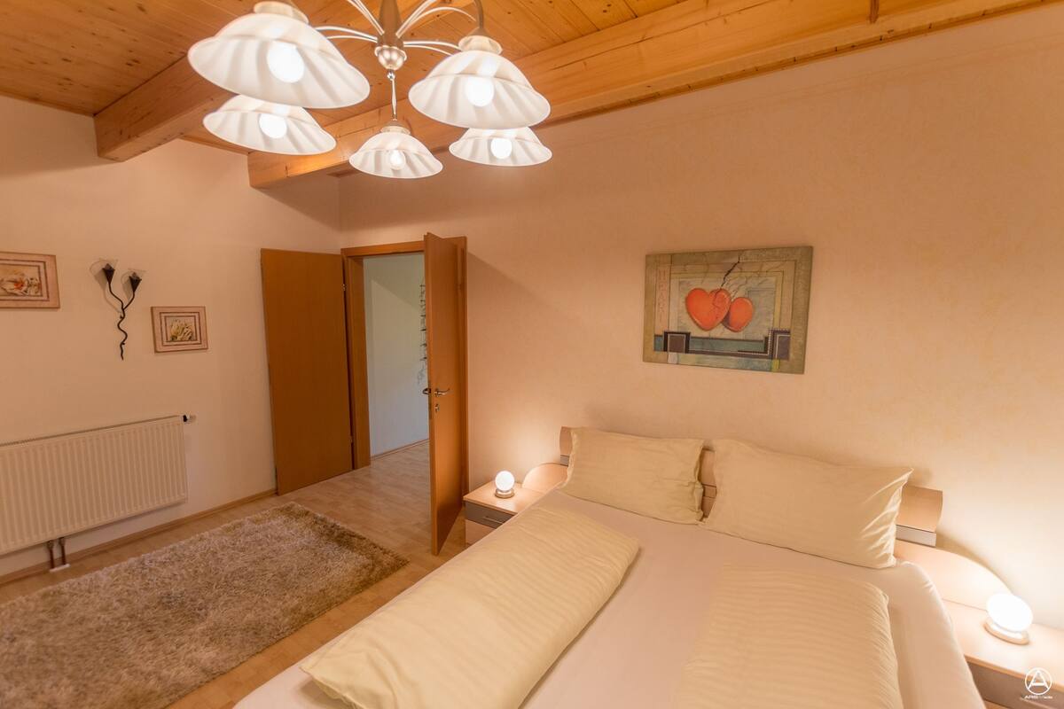 A bedroom with a wooden ceiling, a bed with white bedding, a nightstand with a lamp, a radiator, and a door leading to another room.