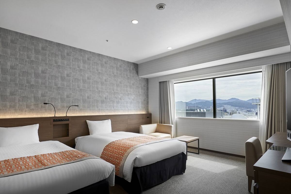 A hotel room with two beds, a desk, a chair, a small table, and a large window offering a city view.