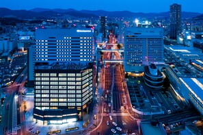 Un paisaje urbano moderno al atardecer con edificios iluminados, un cruce de calles concurrido y un hotel destacado con un letrero.