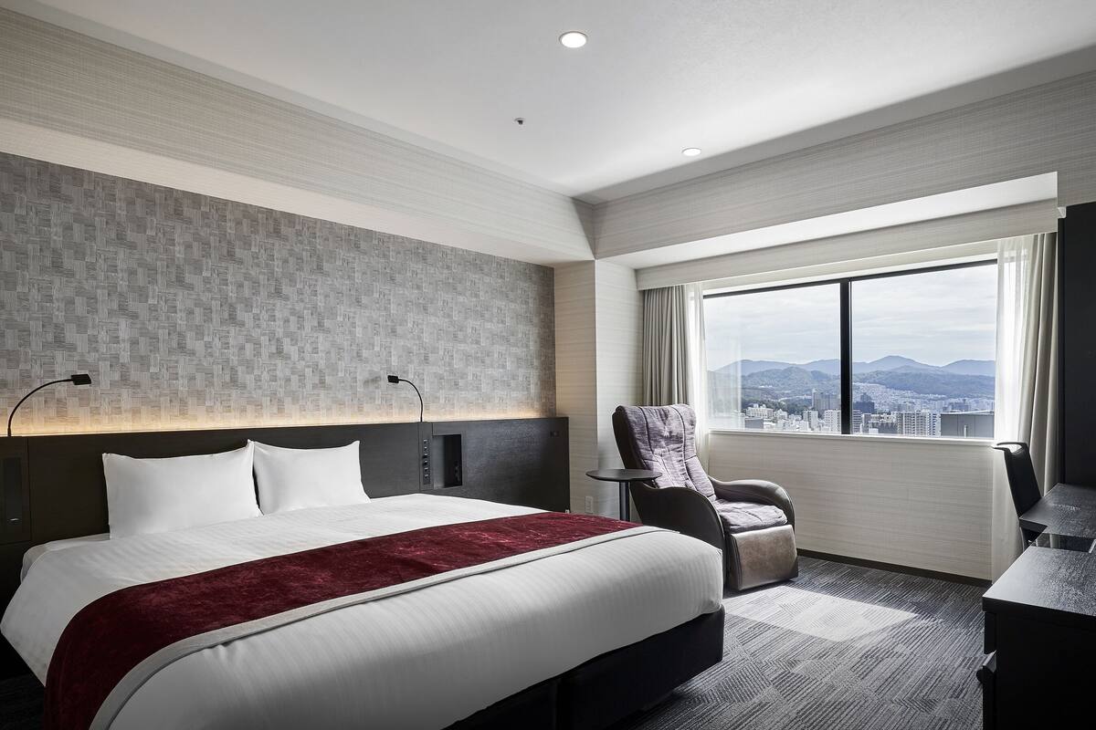 A hotel room with a large bed, a desk, a chair, and a view of the city through the window.