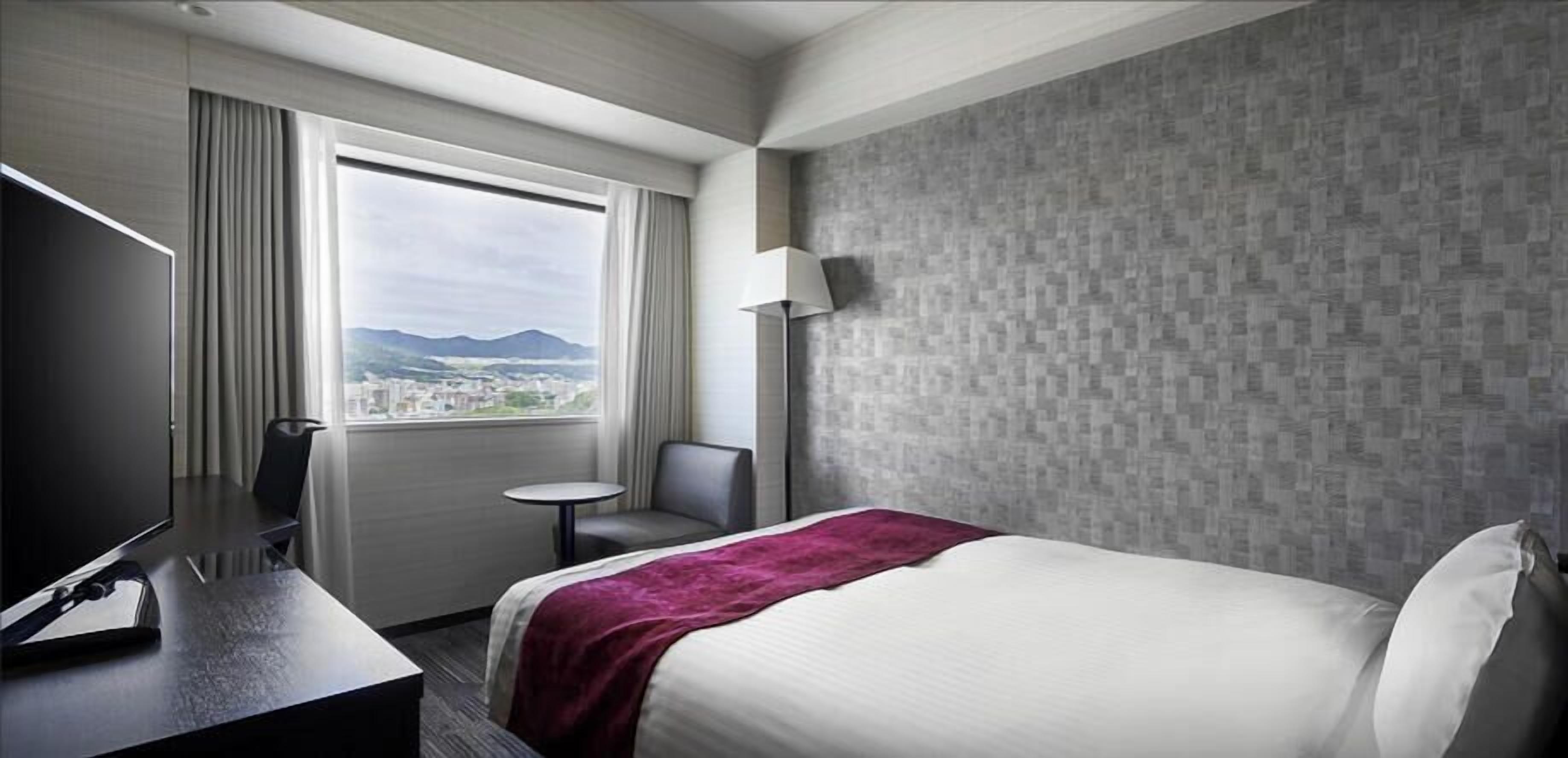A hotel room with a bed, a desk, a chair, a lamp, and a window with a view.