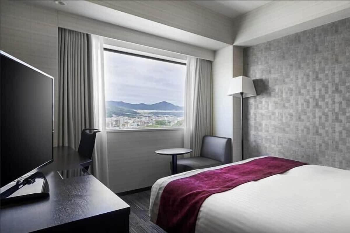 A hotel room with a bed, a desk, a chair, a lamp, and a window with a view.