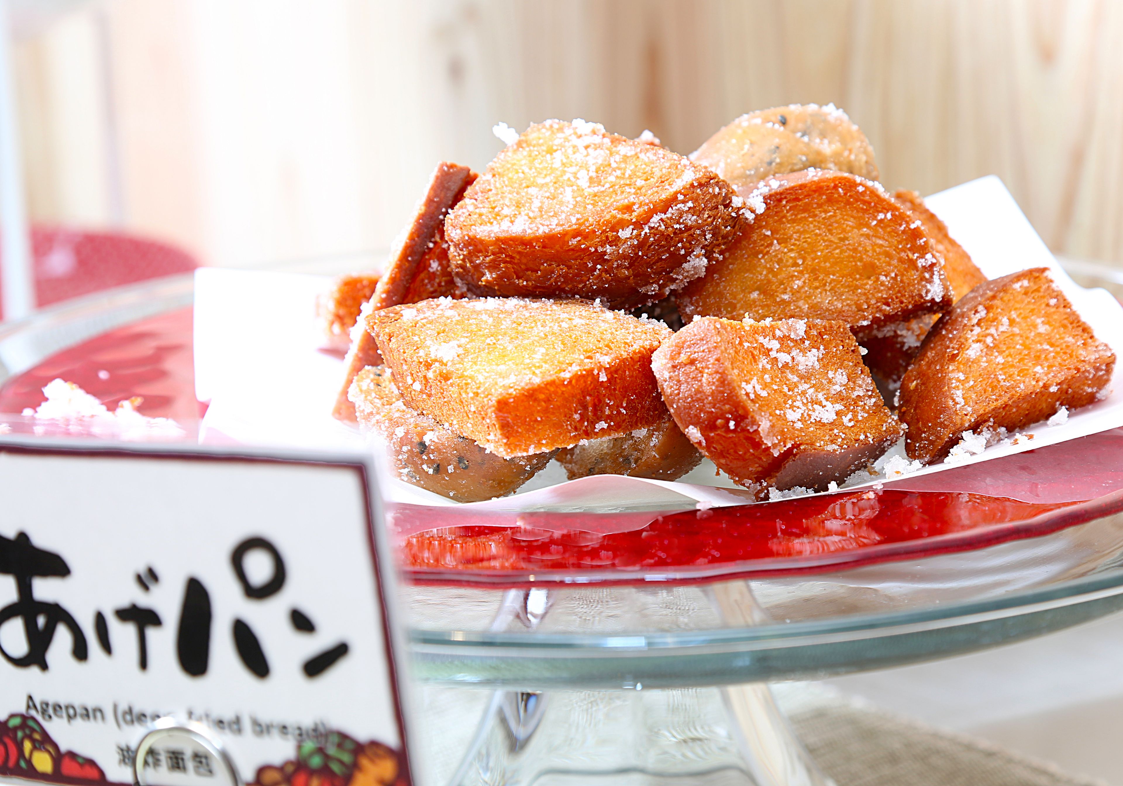 A glass bowl filled with deep-fried bread pieces, dusted with sugar, on a table with a sign in a foreign language.