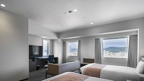 Habitación de hotel con dos camas, un escritorio, una televisión y grandes ventanales con vistas a la ciudad.