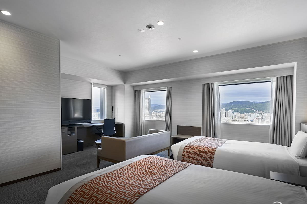 A hotel room with two beds, a desk, a TV, and large windows offering a city view.