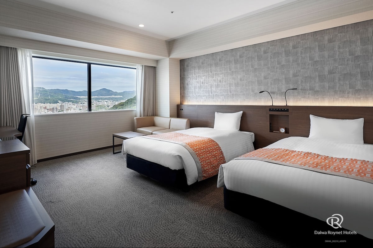 A hotel room with two beds, a desk, and a chair. There is a large window with a view of a city and mountains.