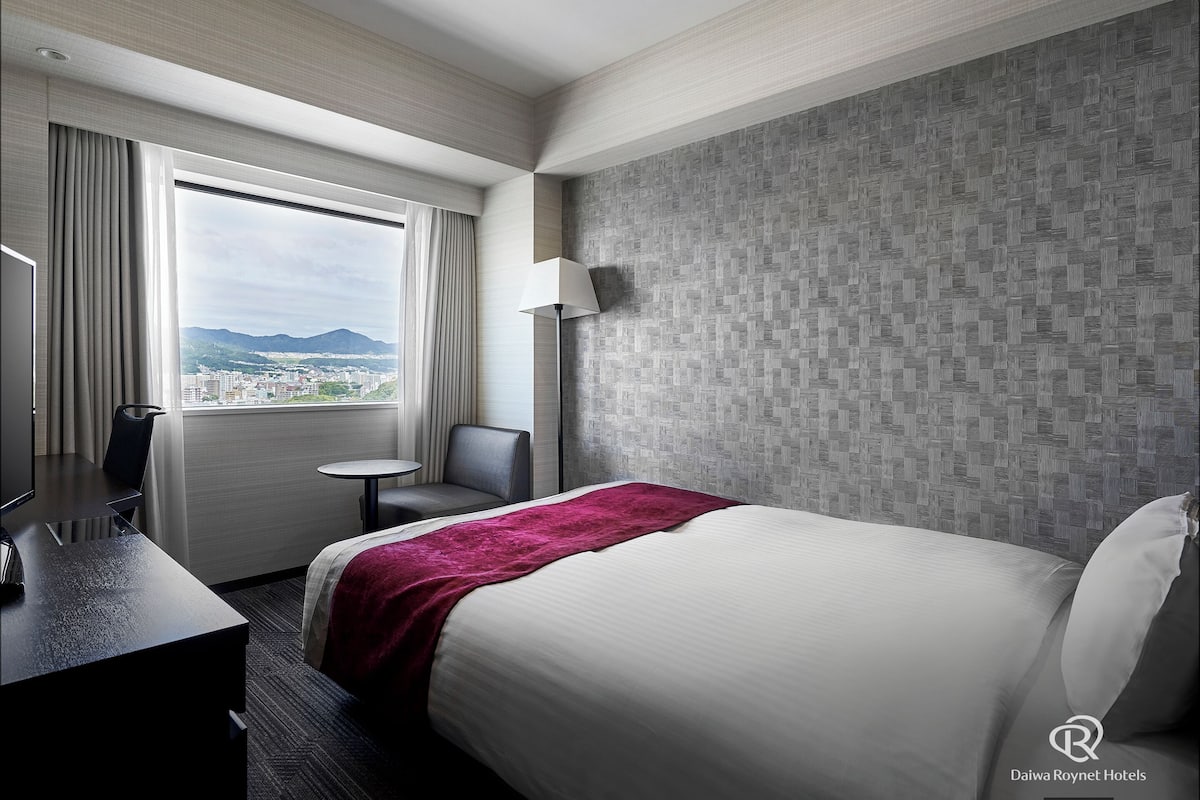A hotel room with a bed, a desk, a chair, a lamp, and a view of a cityscape through the window.