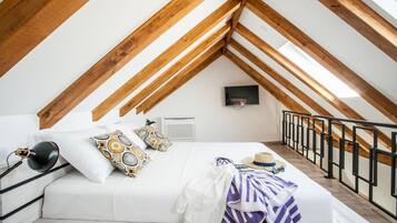 Un dormitorio con techo inclinado, vigas de madera, una cama con almohadones estampados, una mesita de noche con lámpara y un televisor montado en la pared.