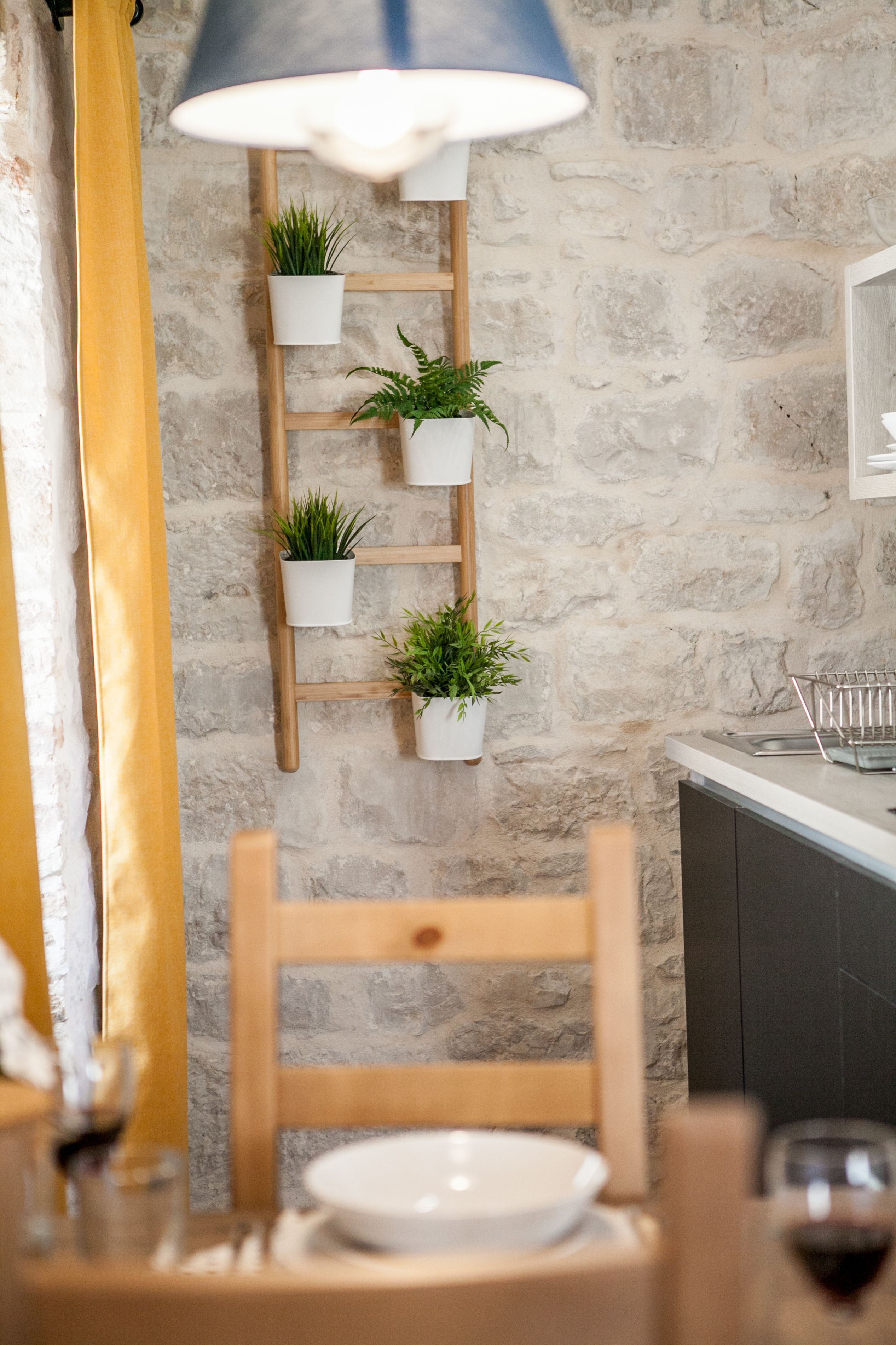 Zona pranzo con una sedia in legno, un tavolo con un piatto bianco e una mensola sospesa con piante, il tutto su un muro in pietra.