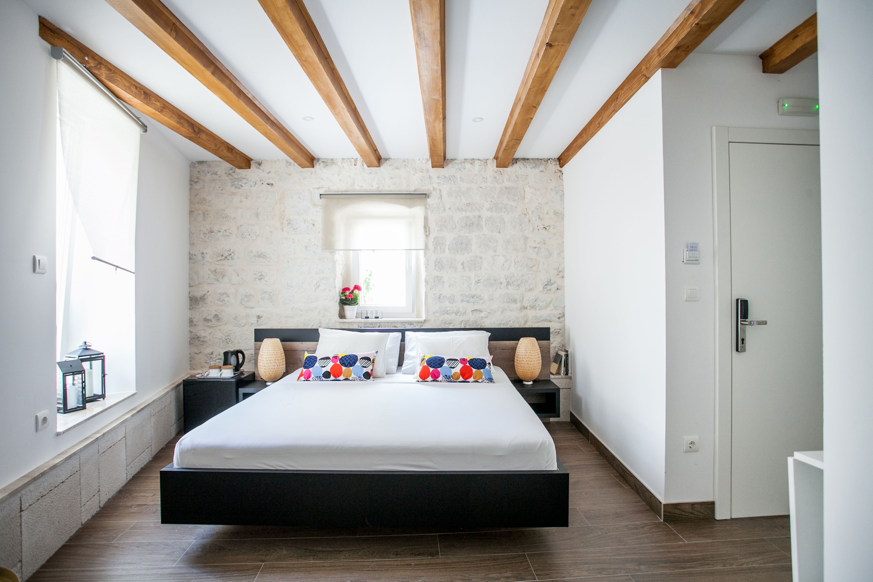 A bedroom with a large bed, wooden beams, stone walls, a window, and a door.