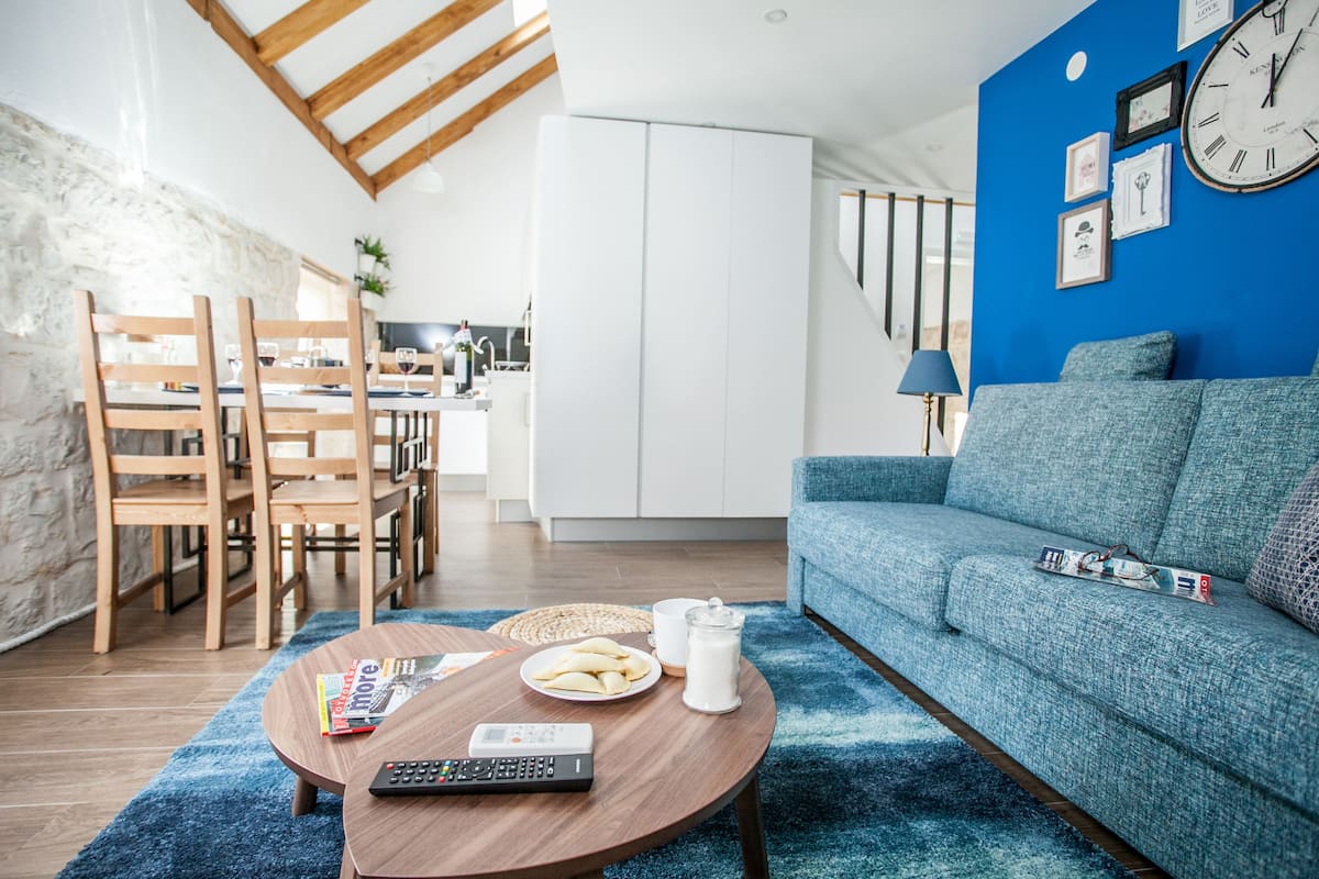 A modern living room with a blue sofa, wooden chairs, a coffee table with remote controls, a magazine, and a plate of pastries.