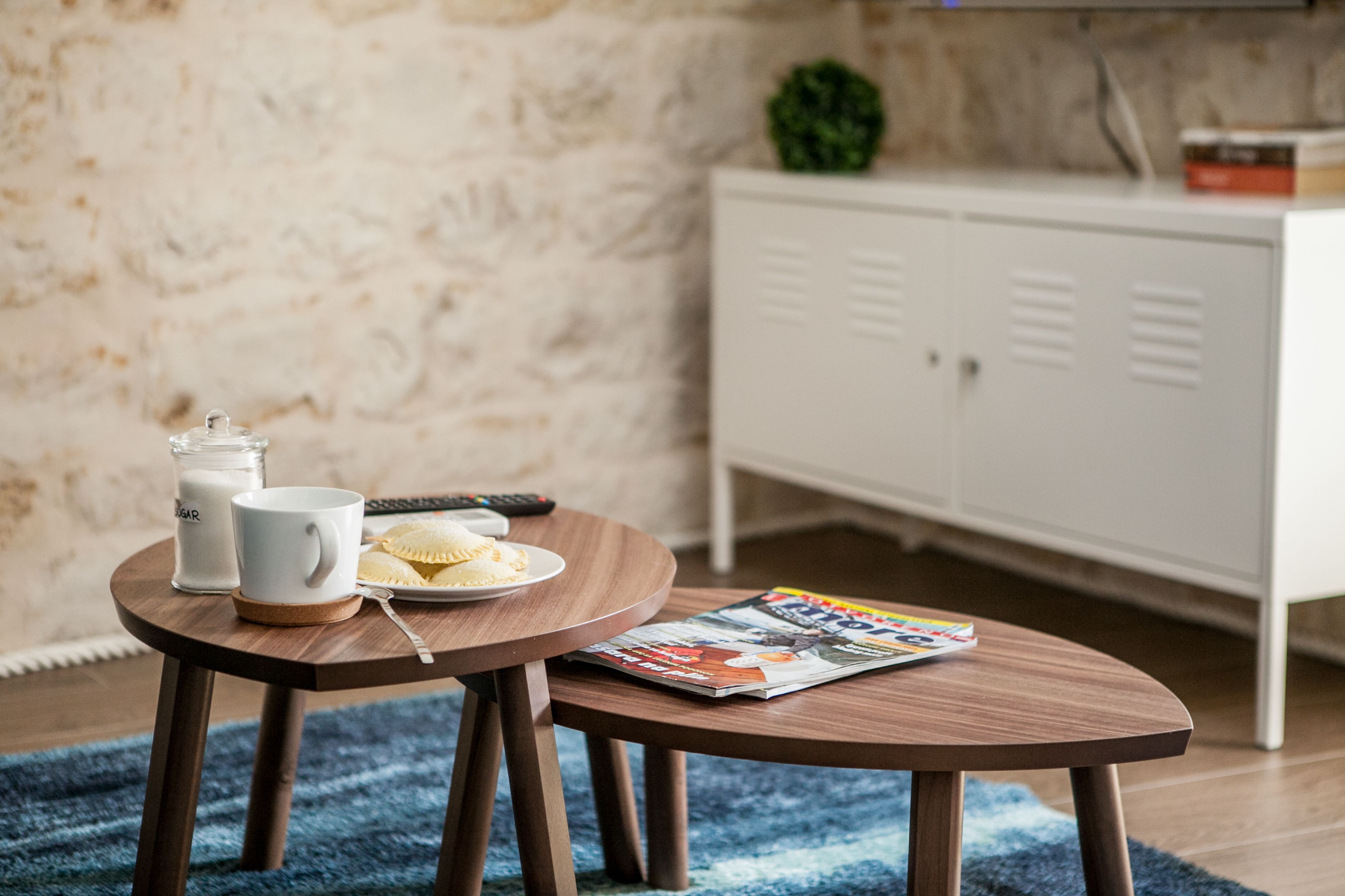 A small round wooden table with a magazine, a glass of milk, a plate with a pastry, and a remote control.