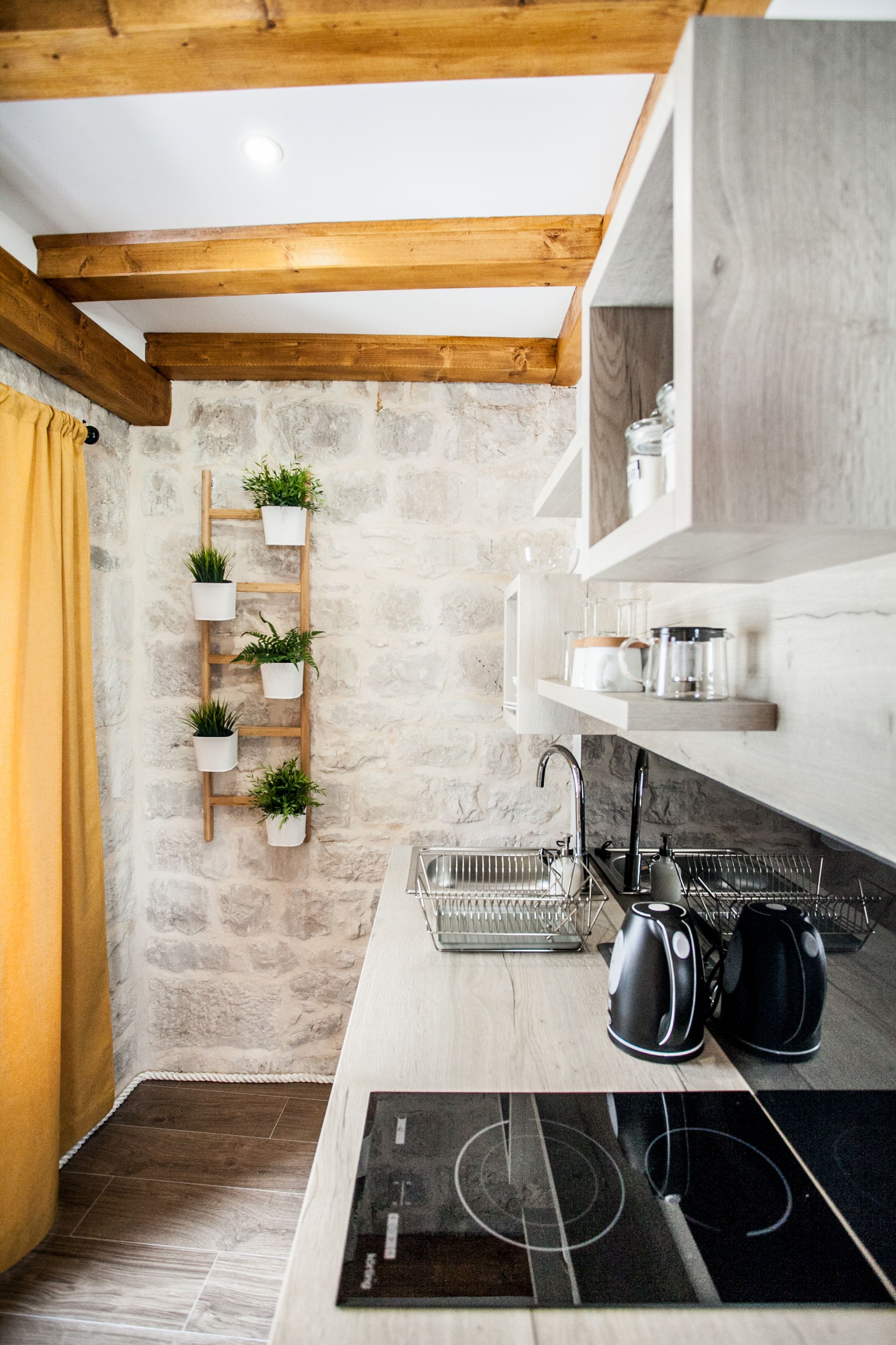 Una cocina con pared de piedra, vigas de madera, una estufa negra y una escalera de madera con plantas en macetas.