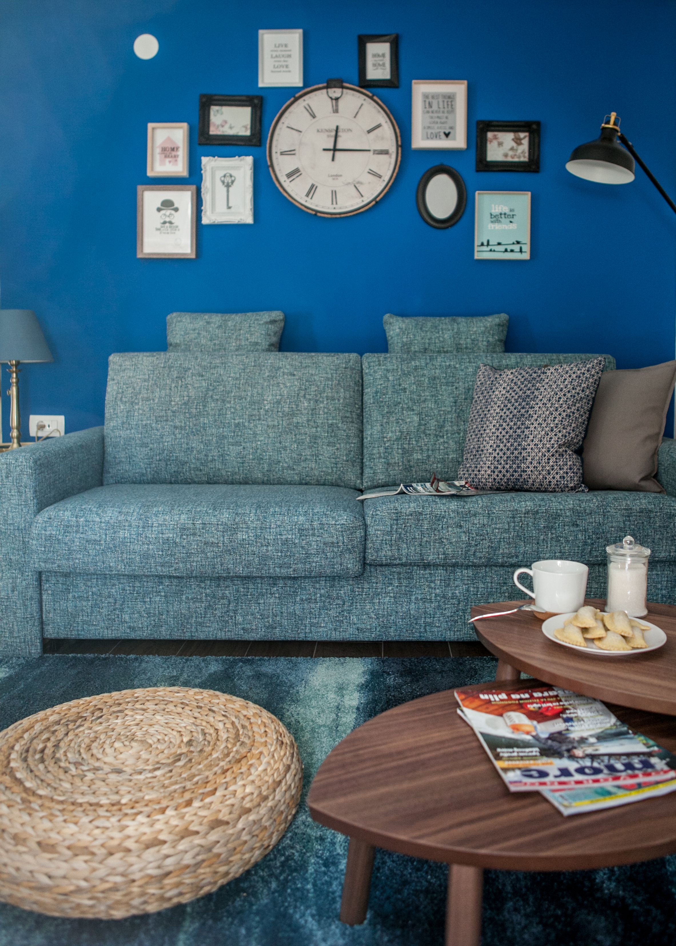 Ein Wohnzimmer mit blauer Wand, einer runden Uhr, gerahmten Bildern, einem Sofa, einem Couchtisch, einer Lampe und einem gewebten Korb.