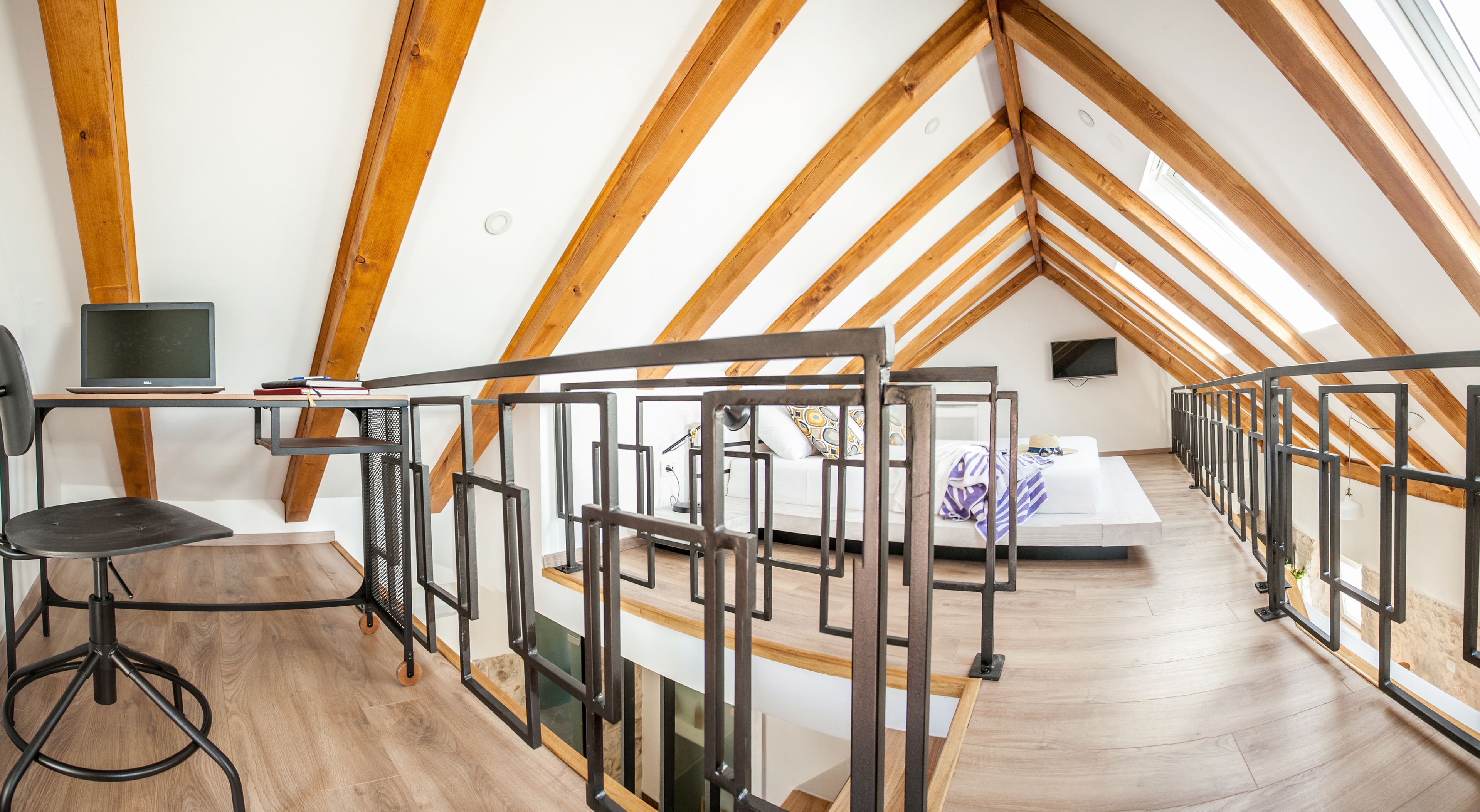 A high-ceilinged attic room with exposed wooden beams, a desk with a laptop, a bed with a patterned bedspread, and a view of the sky through the sloped ceiling.