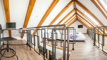 A high-ceilinged attic room with exposed wooden beams, a desk with a laptop, a bed with a patterned bedspread, and a view of the sky through the sloped ceiling.